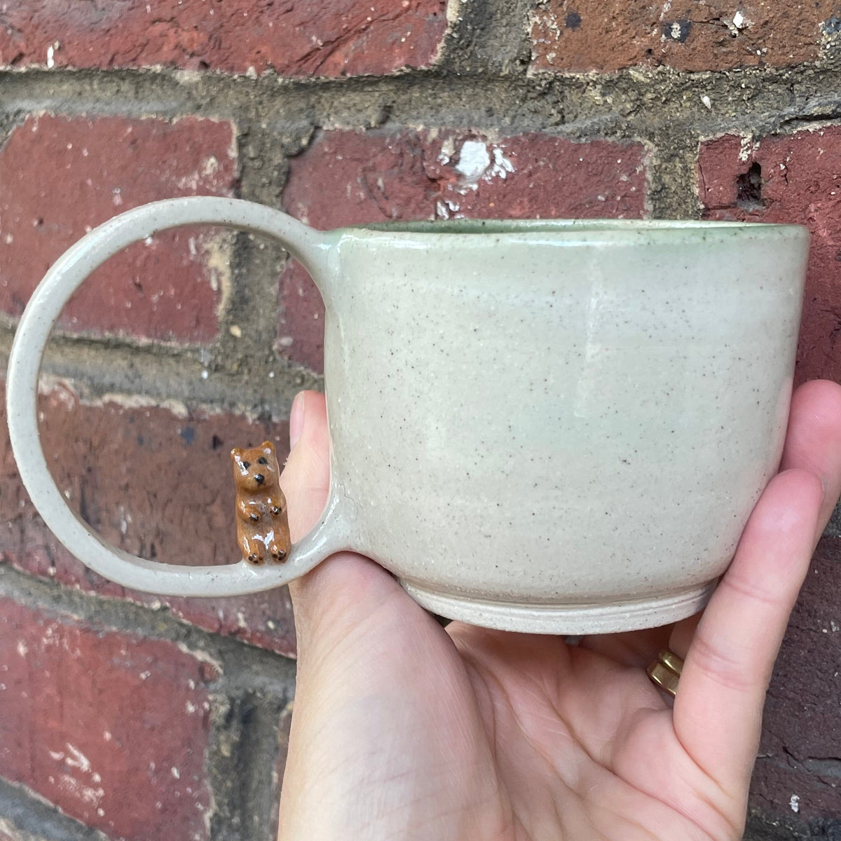 A speckled beige ceramic mug with a loop handle with a bear on it, set against a red brick wall background.